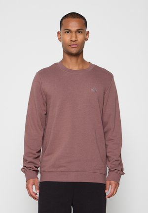 Homme debout, face à l'avant, portant un sweat-shirt mauve à manches longues avec un petit logo sur la poitrine et un pantalon noir.
