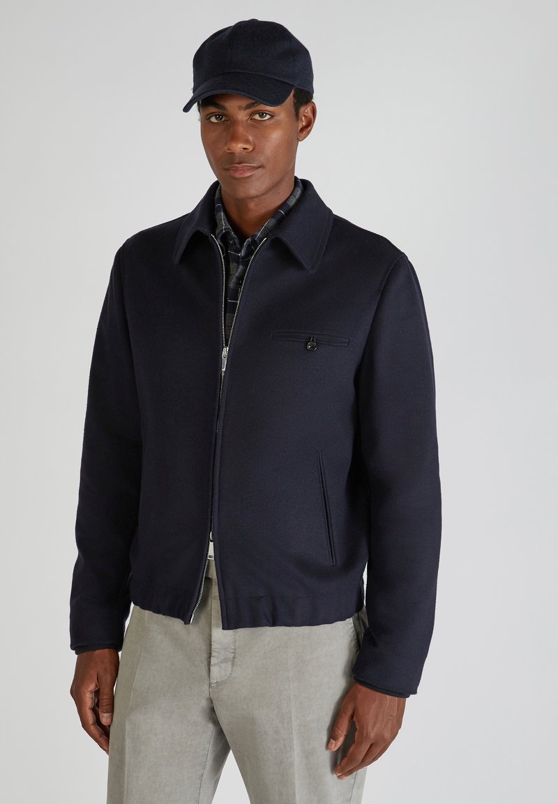 Marineblaues Wollmischjackett mit Kragen, Reißverschluss und einer Brusttasche. Getragen über einem karierter Hemd und kombiniert mit hellgrauen Hosen.