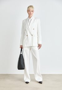 Blazer doppiopetto in lino bianco e pantaloni coordinati, abbinati a scarpe nere con punta a contrasto e una borsa tote nera. Texture liscia e design su misura.