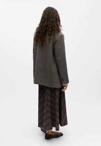 Brauner Wollblazer mit strukturiertem Schnitt, kombiniert mit einem langen, gemusterten braunen Rock mit diagonalen Linien. Schwarze Ballerinas runden das Outfit ab.