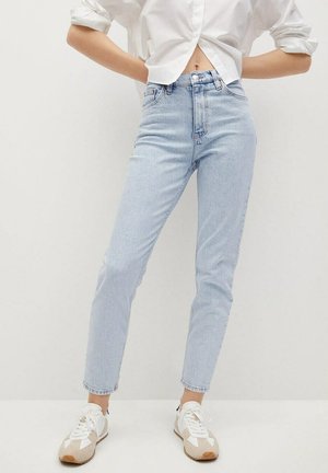 Person trägt hellblaue High-Waist-Jeans, weiße Sneaker und ein weißes, kurz geschnittenes Hemd mit hochgekrempelten Ärmeln.