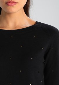 Suéter de punto negro con cuello redondo, con acentos de lentejuelas doradas distribuidos de manera uniforme por la tela. Textura suave, ligero.