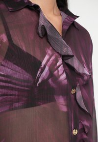 Blusa trasparente viola con colletto a volant, caratterizzata da un motivo di strisce viola piu scure e bottoni dorati. Tessuto morbido, leggero.