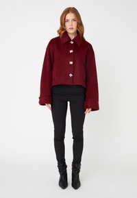 Burgundy cropped jacket with large collar and five decorative buttons; soft texture, wide sleeves, worn over black fitted pants and heeled boots.