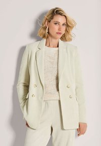 Vrouw met kort blond haar, gekleed in een crèmekleurige corduroy blazer en broek over een gebreide beige top, staand tegen een effen lichte achtergrond.