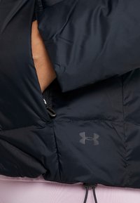 Kurtka puchowa z ciemnogranatowego materiału, z czarnym zamkiem błyskawicznym, logo Under Armour oraz dopasowanym krojem z elastycznymi akcentami przy krawędzi.