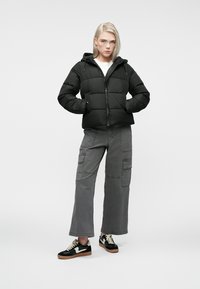 Black puffer jacket with a zip front, hood, and quilted panels, paired with gray cargo pants and black sneakers with white accents.