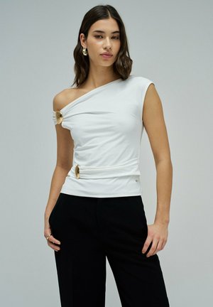 Mujer con top blanco de un solo hombro con broches decorativos dorados y pantalones negros, posando frente a un fondo gris liso.