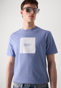 T-shirt de manga curta lilás claro com um patch branco quadrado com o logótipo "BARBOUR INTERNATIONAL". O tecido parece suave, com um ajuste casual.
