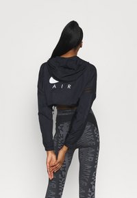 Kort svart vindjacka med huva, med "AIR"-logotyp på ryggen, i kombination med svarta och grå mönstrade leggings. Slät textur.