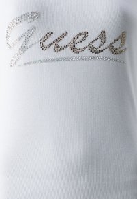 Camiseta de tirantes blanca con canalé que presenta un logo "Guess" que brilla en cristales de imitación, añadiendo textura e interés visual al diseño.