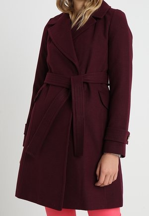 Kvinde iført en knælængde, bæltebåret bordeauxfrakke med reverskraver, klaplommer og knapstropper ved ærmekanterne foran en ensfarvet baggrund.