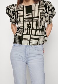 Blusa de manga corta, estampada en beige y negro. Presenta mangas abullonadas, diseños geométricos y un dobladillo recto. Combinada con vaqueros de color azul claro.