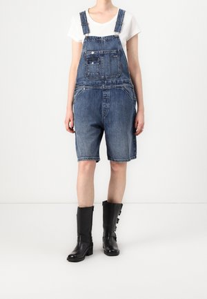 Personne portant une salopette en denim bleu mi-longue, une chemise blanche à manches courtes et des bottes en cuir noires mi-mollet, debout devant un fond blanc uni.