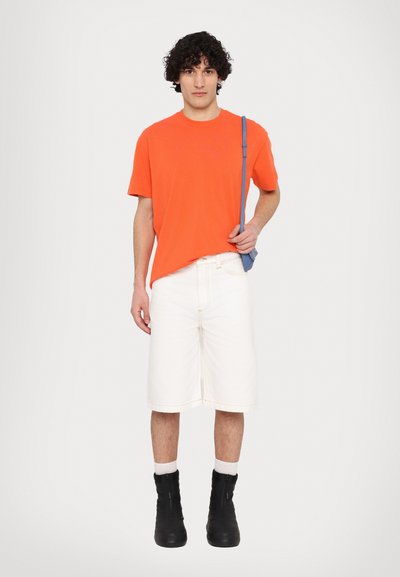T-shirt arancione brillante, pantaloni corto in denim bianchi, stivali neri con zip e borsa a tracolla blu. Il tessuto appare liscio con dettagli minimi.