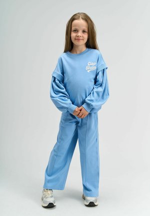 Vaaleansininen huppari, jossa on puffattuja hihoja ja teksti "Ciao Bella". Asua täydentävät löysät housut ja hopeiset tennarit.