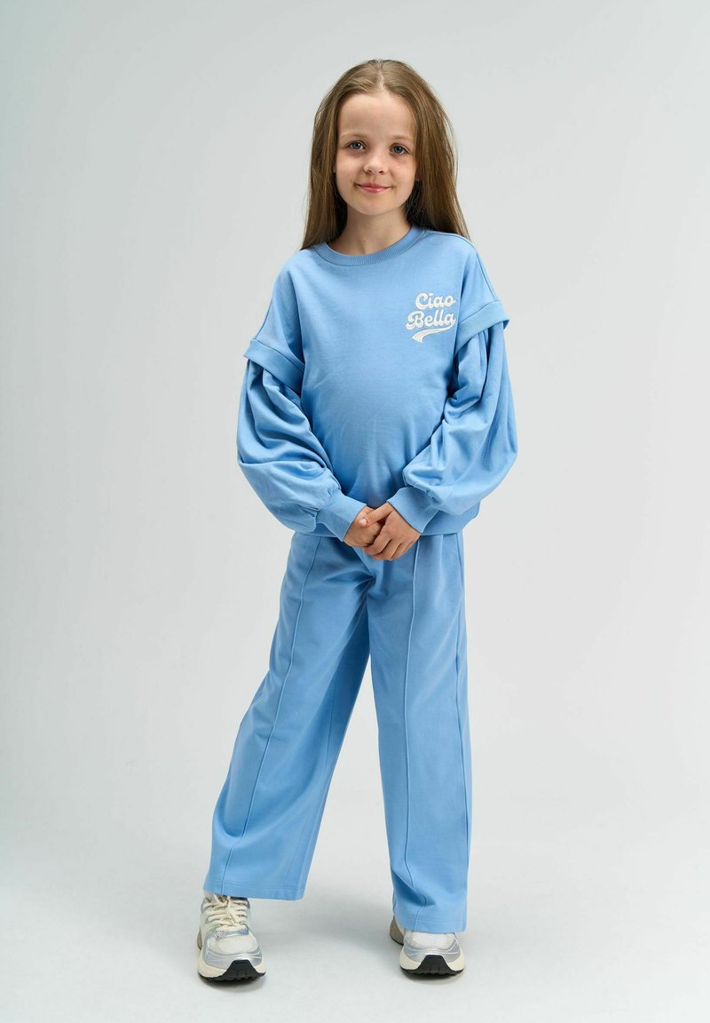 Sweatshirt bleu clair avec des manches bouffantes et le texte "Ciao Bella", accompagné de pantalons amples assortis, complété par des baskets argentées.