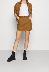 Jupe en coton marron à taille haute, associée à une veste marron zippée et des bottes de combat noires à lacets. Chaussettes blanches visibles au-dessus des bottes.