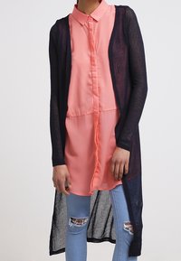 Marinblå lång kofta över en rosa skjorta med knappar. Skjortan har krage och är genomskinlig i nederkant. Ljusblå slitna jeans syns.