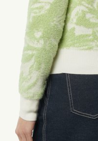 Zielony, puszysty sweter z abstrakcyjnym wzorem, z kremowym ściągaczem u dołu i przy rękawach. Tkanina wydaje się miękka i fakturowana, doskonale komponując się z ciemnym dżinsami.