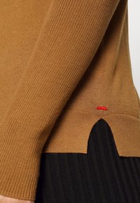 Sweter dziergany w kolorze brązowym, z prążkowanym wzorem, bocznym rozcięciem z falistym wykończeniem i małym czerwonym akcentem w postaci ściegu. Czarny materiał pod spodem.