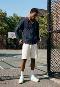 Jeune homme en sweat à capuche bleu marine, short blanc et baskets blanches debout sur un terrain de basket extérieur à côté d'une clôture en grillage.
