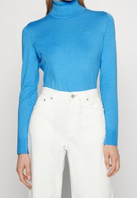 Pull à col roulé bleu en tissu tricoté avec des poignets côtelés, associé à un jean en denim blanc taille haute avec des boutons et des détails de poches.