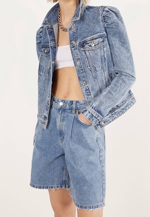 Frau trägt eine hellblaue Acid-Wash-Denimjacke mit aufgepufften Schultern und passende hoch taillierte Denim-Shorts, weißes Croptop und Kettenhalskette.