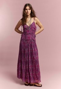 Joven mujer de pie con las manos detrás de la espalda, vistiendo un vestido maxi sin mangas de color púrpura con estampado y collares plateados en capas.