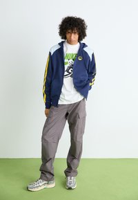 Navy zip-up hoodie with light blue and yellow accents, paired with a white Nike graphic t-shirt and gray cargo pants. Chunky sneakers complete the look.