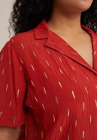 Blusa roja hecha de un tejido texturizado con un aspecto arrugado y acentos metálicos dorados en un patrón simétrico y alargado. Mangas cortas y cuello.