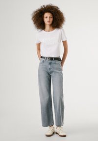 Hvit t-skjorte med "pepe jeans" trykk, parret med lyse, vide jeans og et svart belte. Modellen har på seg beige joggesko.