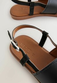 Sandali in pelle nera con cinturini regolabili, dotati di una soletta imbottita marrone e suola piatta. Dettaglio logo inciso sulla soletta.
