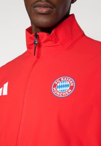 Red jacket featuring a zip closure, high collar, and FC Bayern München logo. Crafted from smooth fabric with white accent on the sleeve.
