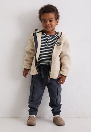 Crèmekleurig fleecejack met marineblauwe accenten, gestreept overhemd met knopen, grijze cargobroek en beige schoenen. Voorzien van zakken en elastische manchetten.