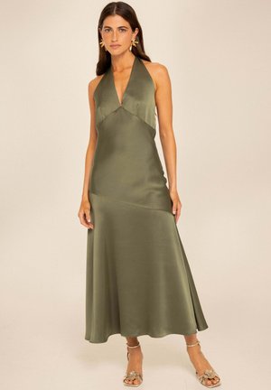 Mujer con un vestido midi de tirantes color verde oliva con escote en V estilo halter, pendientes florales dorados y sandalias.