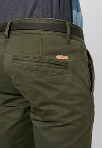 Zelené bavlněné kalhoty s hladkou texturou, vybavené černým opaskem a mosazným logem EDC na zadní kapse.