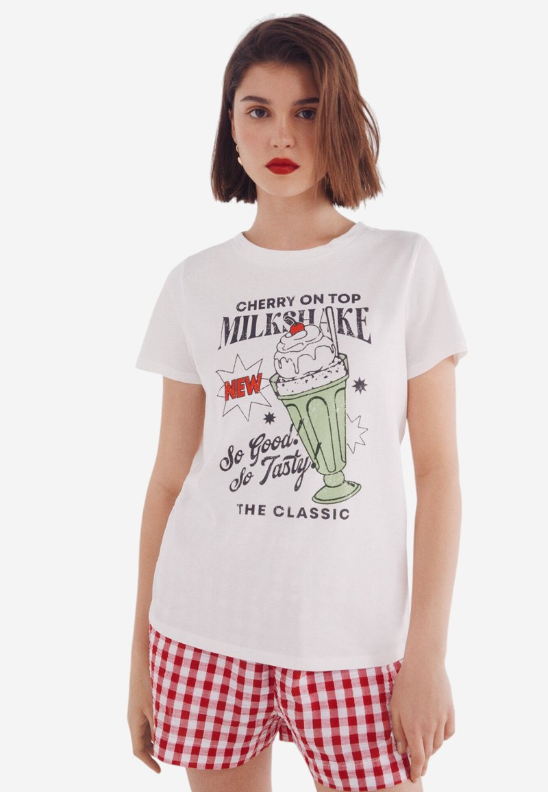 Witte katoenen T-shirt met een kleurrijke milkshake-afbeelding, voorzien van een kers bovenop en tekst. Gecombineerd met rood-wit geruite shorts.