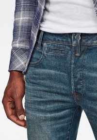 Jeans de mezclilla azul oscuro con una textura suave, costuras visibles y un bolsillo delantero. Combinados con una camisa de cuadros en azul y blanco.