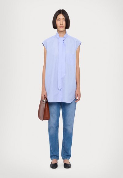 Blusa senza maniche celeste con dettaglio a nodo, abbinata a jeans celesti e scarpe nere slip-on, mentre tiene una borsa marrone.