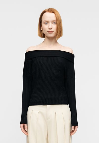 Pull noir épaules dénudées avec texture côtelée, manches longues et motif diagonal ; associé à un pantalon plissé beige.