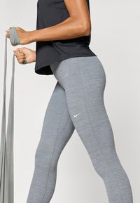 Γκρι αθλητικό κολάν με υφή και αντίθετο λευκό λογότυπο Nike, συνδυασμένο με μαύρο εφαρμοστό τοπ και κορδέλα γυμναστικής στο χέρι.