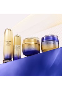 Shiseido Vital Perfection Hautpflegesammlung mit vier Produkten in Glasbehältern mit goldenen und blauen Farbverläufen, platziert auf einer blauen Oberfläche.