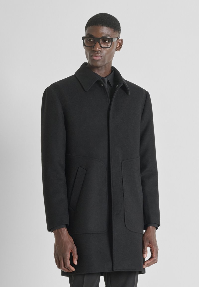 Cappotto in misto lana nero con colletto a punta, due tasche frontali e una silhouette snella. Presenta una chiusura con bottoni e un tessuto testurizzato.