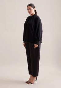 Schwarze Langarmbluse mit gerafftem Ausschnitt und knöpfbaren Manschetten, kombiniert mit schwarzen, maßgeschneiderten Hosen und schwarzen Sandalen mit hohen Absätzen.