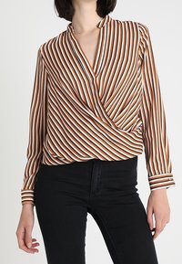 Blouse portefeuille à manches longues avec un motif rayé blanc, orange et marron. Elle présente un col en V profond et un tissu texturé, associée à un pantalon noir.