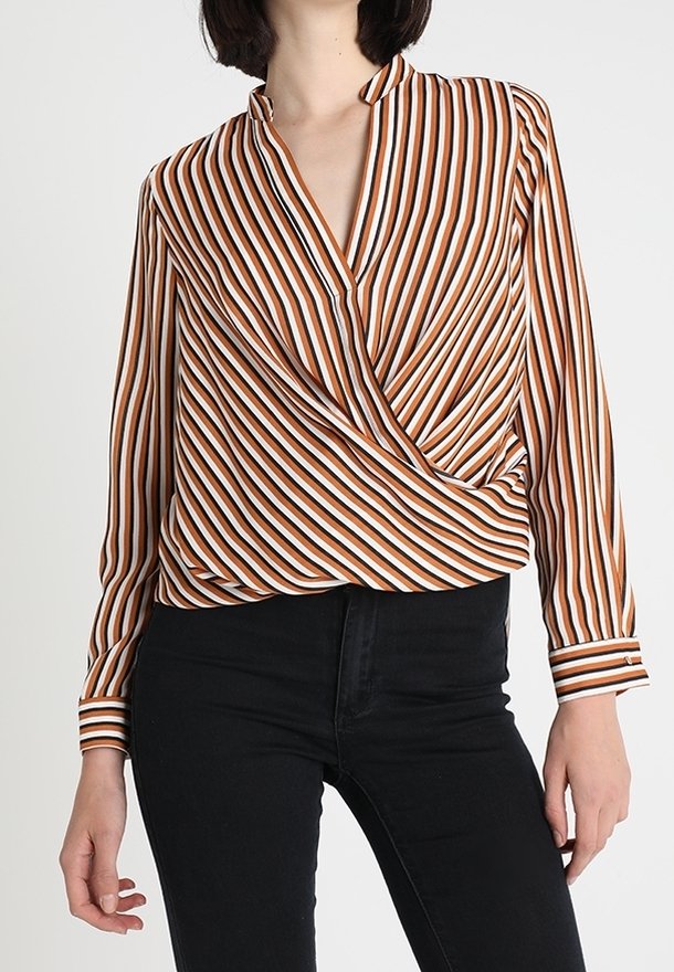 Blouse portefeuille à manches longues avec un motif rayé blanc, orange et marron. Elle présente un col en V profond et un tissu texturé, associée à un pantalon noir.