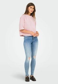 Kvinde stående iført en lys pink oversized t-shirt, slidte lyseblå skinny jeans og mørkebrun loafers på en ensfarvet baggrund.
