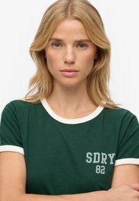Groen T-shirt met korte mouwen, voorzien van een witte geribbelde kraag en mouwomslagen, met een grijze bedrukte logo "SDRY 82" aan de linkerkant.