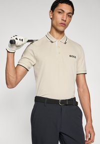 Bézs színű galléros póló kontrasztos fekete szegéllyel, amelynek mellrészén logó található. Sötét nadrággal és fonott fekete övvel párosítva.